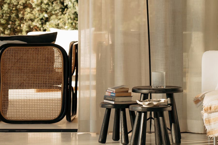 Rattan armchair beside sheer curtains, with nested stools styled with books and a candle