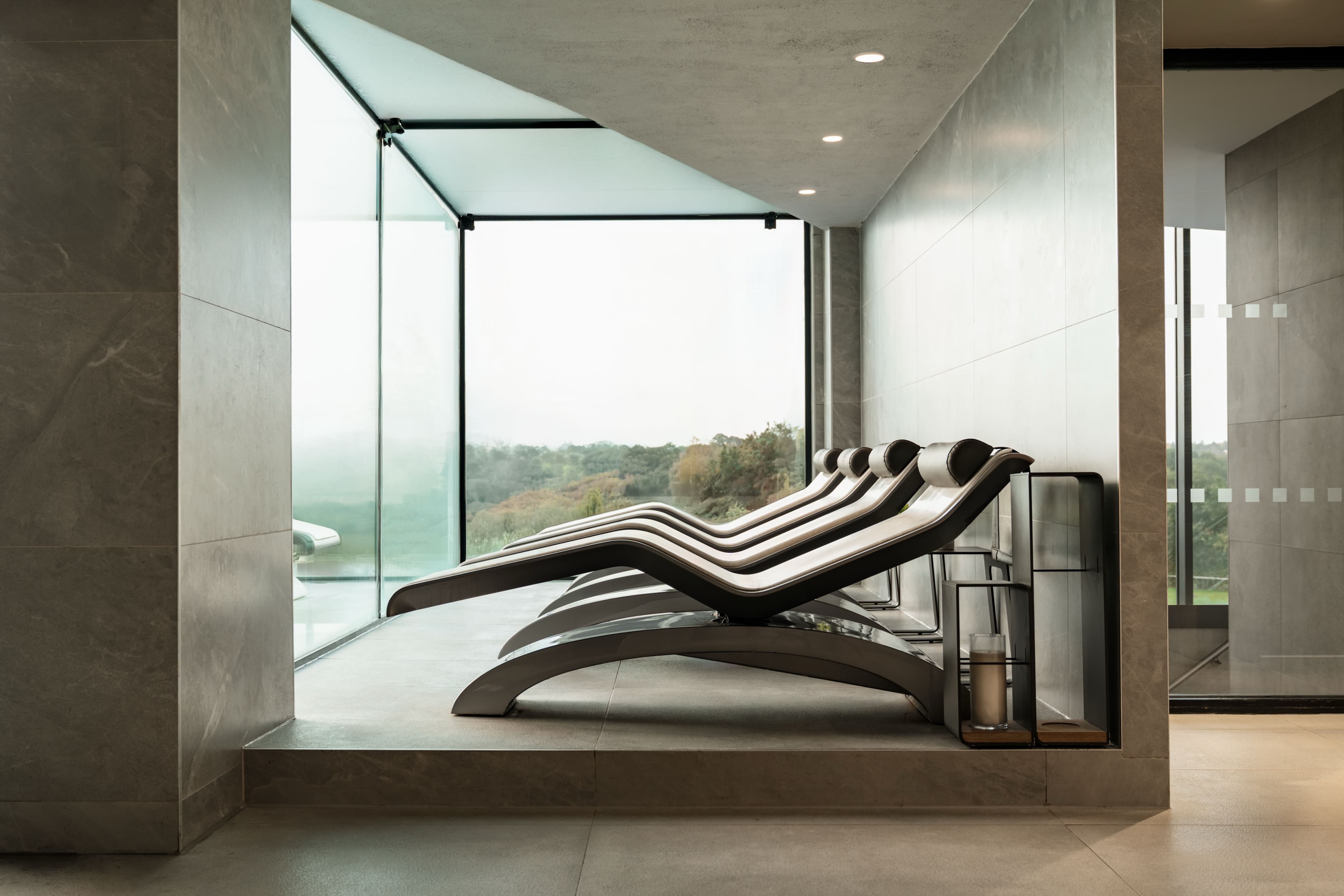 Row of curved heated loungers in a stone-clad spa alcove with floor-to-ceiling windows onto trees